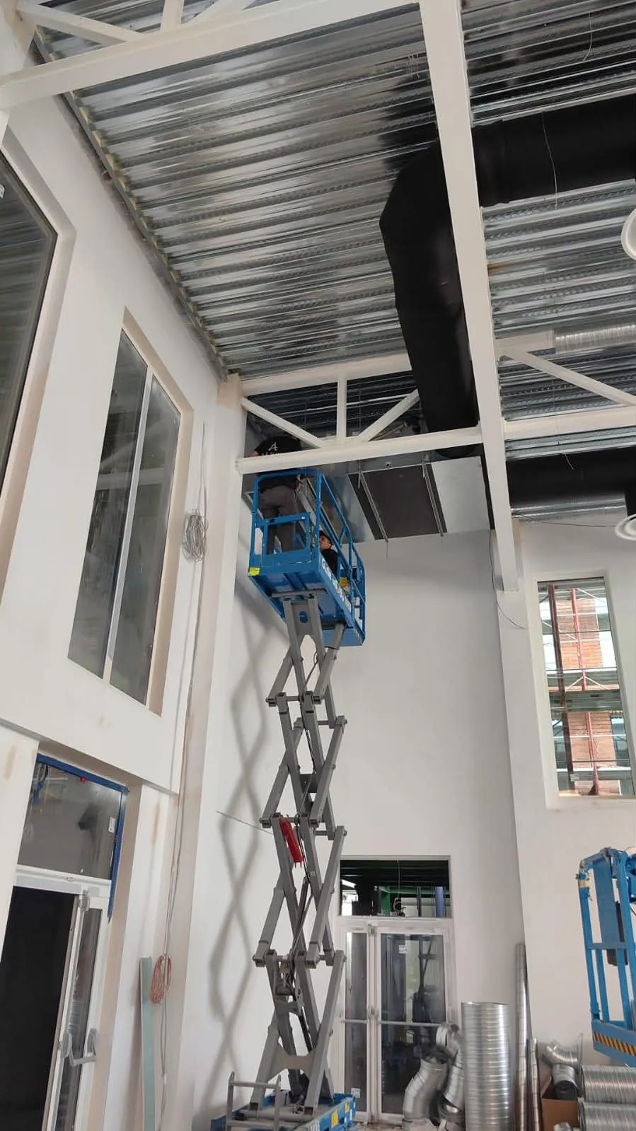 Un lucrător pe o platformă elevatoare albastră lucrează la o unitate montată sub grinzile unei hale înalte, lângă o tubulatură învelită în izolație neagră